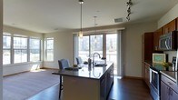 This single-floor space showcases a thoughtfully designed open-concept layout that seamlessly connects the kitchen and living areas. The kitchen serves as the centerpiece, anchored by a substantial island topped with dark granite countertops and an undermount sink, offering both functionality and visual appeal. Sleek pendant lights hang above the island, complementing a track lighting system that adds a modern, versatile touch to the space. The cabinetry and dark hardwood flooring throughout contribute to a sophisticated, contemporary aesthetic. Large windows and a sliding glass door flood the interior with abundant natural light while framing views of the surrounding neighborhood. The neutral color palette, featuring soft white walls and warm wood tones, creates a calm and inviting ambiance. Stylish upholstered bar stools along the island provide casual seating, making the kitchen a natural gathering spot. The open flow between the kitchen and adjacent living area enhances the sense of spaciousness, making this a well-appointed and practical space suited for both everyday living and entertaining.