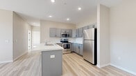 This single-floor residence offers a thoughtfully designed open-concept layout that seamlessly connects the kitchen and living areas. The kitchen is a standout feature, showcasing grey cabinetry, a central peninsula with a white countertop and built-in sink, and a full suite of stainless steel appliances including a refrigerator, electric range, microwave, and dishwasher. Light wood-look luxury vinyl flooring runs throughout the main living areas, complementing the neutral warm-beige wall tones and recessed lighting overhead. The open living area is bright and airy, benefiting from natural light through a large window and a ceiling fan for added comfort. The bedrooms are carpeted and well-lit, each equipped with ceiling fans and windows that allow generous natural light to fill the space. A clean, functional bathroom features wood-look flooring, neutral walls, and matte black hardware accents. The attached garage offers generous vehicle and storage space with drywall-finished walls and an automatic opener. The home presents a fresh, modern aesthetic with consistent finishes throughout, making it a comfortable and well-appointed single-level living space.