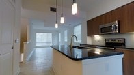 This contemporary multi-floor space spans three levels and is designed with a clean, modern aesthetic throughout. The open-concept main living area seamlessly connects the kitchen and living space, creating a fluid and functional layout ideal for everyday living and entertaining.

The kitchen is a standout feature, anchored by a large island with striking dark granite countertops and an elegant gold-finish faucet. Warm wood cabinetry contrasts beautifully against crisp white walls and light-toned tile flooring. A trio of pendant lights hangs above the island, providing focused task lighting while adding a stylish architectural touch. Built-in stainless steel appliances, including a microwave, are neatly integrated into the cabinetry for a streamlined look.

The adjacent living area extends naturally from the kitchen, bathed in abundant natural light streaming through multiple windows dressed with horizontal blinds. The consistent tile flooring flows throughout, reinforcing the cohesive, open feel of the space. White walls and high ceilings amplify the sense of volume and brightness across the main level.

With three floors offering ample room to spread across, this space combines thoughtful design, quality finishes, and a bright, welcoming atmosphere that makes it both practical and visually appealing.