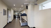 This fitness center is designed as a long, narrow space with a clean, functional layout that maximizes the available floor area. The interior features neutral, light-toned walls paired with dark rubber flooring, creating a practical and professional environment suited for regular exercise use.

Recessed lighting runs the length of the ceiling, ensuring consistent illumination throughout the space. Small horizontal windows are positioned along one wall, allowing modest natural light to supplement the overhead lighting. A wall-mounted monitor and speaker system provide entertainment and audio options during workouts.

The cardio section is prominently featured along the main corridor, housing a treadmill, elliptical trainer, and stationary bike from Vision Fitness, arranged in a row to allow easy access and movement between machines. Further into the space, a dedicated strength training area accommodates a multi-use cable machine, adjustable weight bench, leg press equipment, and a full dumbbell rack with weights neatly organized by size.

The far end of the facility opens into a slightly wider workout area with additional free-weight equipment and mirrors that visually expand the space. Two access doors provide entry and exit points. A fire extinguisher and exit signage are clearly visible, ensuring safety compliance throughout the facility.