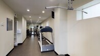 This fitness center is designed as a long, narrow space with a clean, functional layout that maximizes the available floor area. The interior features neutral, light-toned walls paired with dark rubber flooring, creating a practical and professional environment suited for regular exercise use.

Recessed lighting runs the length of the ceiling, ensuring consistent illumination throughout the space. Small horizontal windows are positioned along one wall, allowing modest natural light to supplement the overhead lighting. A wall-mounted monitor and speaker system provide entertainment and audio options during workouts.

The cardio section is prominently featured along the main corridor, housing a treadmill, elliptical trainer, and stationary bike from Vision Fitness, arranged in a row to allow easy access and movement between machines. Further into the space, a dedicated strength training area accommodates a multi-use cable machine, adjustable weight bench, leg press equipment, and a full dumbbell rack with weights neatly organized by size.

The far end of the facility opens into a slightly wider workout area with additional free-weight equipment and mirrors that visually expand the space. Two access doors provide entry and exit points. A fire extinguisher and exit signage are clearly visible, ensuring safety compliance throughout the facility.