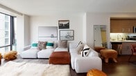 This well-appointed single-floor residence offers a thoughtfully designed open-concept layout that seamlessly integrates the living, dining, and kitchen areas. The living room is a comfortable and stylish space, anchored by a large sectional sofa and accented with curated wall art, creating a welcoming atmosphere. Dark hardwood flooring runs throughout the main living areas, providing a warm, grounding contrast to the crisp white walls.

The kitchen is a standout feature, outfitted with rich wood-grain cabinetry that extends to the ceiling, under-cabinet lighting, a white tile backsplash, and a full suite of stainless steel appliances including a gas range, built-in microwave, dishwasher, and a large refrigerator. The adjacent dining area accommodates a rustic wood dining table with industrial-style seating, perfect for everyday meals or entertaining.

The bedroom is a serene retreat, bathed in natural light from a large window with urban views. The clean, neutral palette and minimalist design give the room a calm, restful ambiance.

The bathroom is finished with light tile flooring, a modern floating vanity with wood-tone cabinetry, a large mirror, and a full-size soaking tub with a shower, offering both functionality and clean contemporary style throughout.
