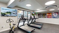 This fitness center is organized across two dedicated exercise rooms, together offering a well-rounded environment for a full-body workout experience. The space features a clean, modern aesthetic with crisp white walls, durable speckled rubber flooring, and recessed circular flush-mount ceiling lights that provide bright, even illumination throughout.

The cardio-focused room is lined with large floor-to-ceiling windows that invite natural light and offer outdoor views, creating an open, energizing atmosphere. Equipment in this area includes multiple treadmills, a stair climber, a stationary bike, elliptical trainers, and a rowing machine — catering to a wide range of cardio preferences. Wall-mounted flat-screen TVs are positioned for easy viewing during workouts, and framed sports jerseys add a spirited decorative touch.

The strength and functional training room features a cable machine, free weights, adjustable benches, and a wall of mirrors that enhance the sense of space. A dedicated functional training rack holds resistance bands, medicine balls, and a foam roller, supporting dynamic and mobility-focused workouts. Additional windows maintain visual connection to the outdoors, keeping the space feeling bright and welcoming. Together, the two rooms provide a comprehensive and thoughtfully designed fitness facility.