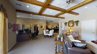 This single-floor space offers a well-appointed, open-plan layout that seamlessly integrates multiple functional areas within one expansive interior. The most striking architectural feature is the coffered ceiling adorned with rich, warm-toned wooden beams, which adds a sense of structure and elegance to the space. Recessed lighting and ceiling fans are integrated throughout, ensuring both comfort and consistent illumination.

The main gathering area is anchored by a plush leather sofa and a classic wooden console table, creating a relaxed yet refined lounge setting. Adjacent to this is a formal dining area furnished with upholstered white chairs and a traditional wooden dining table, ideal for group meals or meetings. Decorative wall mirrors and warm-toned accent lamps contribute to the inviting ambiance.

Along the perimeter, a dedicated desk and office chairs indicate a functional workspace or reception area, making the space versatile for both social and administrative purposes. In the background, a bar-height counter with seating extends the space further, suggesting a café or refreshment area. Neutral carpeting and warm beige walls tie the various zones together, lending a cohesive, comfortable atmosphere throughout this thoughtfully designed communal environment.