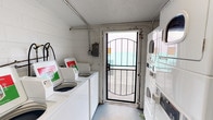 This single-floor laundry facility is a practical, no-frills space designed to serve residents with all essential washing and drying needs. The room features white-painted concrete block walls and a light-colored floor, giving the space a clean and utilitarian aesthetic. Natural light filters in through the glass panel of a black decorative security door, which provides both ventilation and visibility to the exterior.

Along one wall, a row of top-loading washing machines is neatly arranged side by side, each equipped with coin-operated mechanisms and informational signage to guide users. On the opposite side, stacked dryer units maximize vertical space, making efficient use of the compact footprint. An electrical panel is mounted on the wall, and visible utility conduits run along the upper portions of the walls, reflecting the functional nature of the space.

The overall ambiance is bright and straightforward, prioritizing accessibility and practicality. The secure entry door with its arched decorative ironwork adds a subtle architectural detail to an otherwise utilitarian environment. This well-organized laundry room offers residents a convenient, self-service option in a clean and manageable setting.