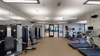 This fitness center is organized into two distinct yet connected zones, offering a comprehensive wellness environment. The main gym area features carpeted flooring and is outfitted with a robust selection of strength-training machines, including cable and resistance equipment, along with cardio machines such as treadmills, ellipticals, and a stair climber. Large wall-mounted mirrors amplify the sense of space, while linear pendant lighting provides bright, even illumination throughout. A wall-mounted television and a distinctive honeycomb-style wooden shelving unit add decorative character to the cardio area, which also benefits from outdoor-facing windows that bring in natural light and views of the surrounding grounds.

The adjacent studio space features light hardwood flooring and an open, unobstructed layout ideal for group fitness classes, yoga, or stretching. A large interactive display screen is mounted on a mirrored wall, enabling guided workouts, while motivational artwork decorates the walls. Glass-paneled doors connect the studio to the gym, maintaining visual continuity. A separate area with wood-look flooring opens directly to an outdoor courtyard through full-length black-framed glass doors, providing a bright, airy setting for stretching benches and rehabilitation-style equipment. The overall design is clean, modern, and functionally well-planned.