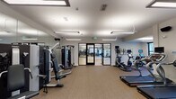 This fitness center is organized into two distinct yet connected zones, offering a comprehensive wellness environment. The main gym area features carpeted flooring and is outfitted with a robust selection of strength-training machines, including cable and resistance equipment, along with cardio machines such as treadmills, ellipticals, and a stair climber. Large wall-mounted mirrors amplify the sense of space, while linear pendant lighting provides bright, even illumination throughout. A wall-mounted television and a distinctive honeycomb-style wooden shelving unit add decorative character to the cardio area, which also benefits from outdoor-facing windows that bring in natural light and views of the surrounding grounds.

The adjacent studio space features light hardwood flooring and an open, unobstructed layout ideal for group fitness classes, yoga, or stretching. A large interactive display screen is mounted on a mirrored wall, enabling guided workouts, while motivational artwork decorates the walls. Glass-paneled doors connect the studio to the gym, maintaining visual continuity. A separate area with wood-look flooring opens directly to an outdoor courtyard through full-length black-framed glass doors, providing a bright, airy setting for stretching benches and rehabilitation-style equipment. The overall design is clean, modern, and functionally well-planned.