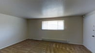 This single-floor space offers a straightforward, functional layout designed for comfortable everyday living. The main living area is finished with wall-to-wall carpet in a neutral tan tone, providing a warm underfoot feel throughout. The ceiling features a textured popcorn finish, adding a classic residential character to the space. A wide, horizontal window fitted with horizontal blinds serves as the room's primary source of natural light, brightening the interior and creating an open, airy atmosphere. Below the window runs a baseboard heating unit, ensuring efficient and consistent warmth throughout the cooler months. The walls are painted in a soft, neutral beige tone that complements the flooring and contributes to the overall understated aesthetic. Electrical outlets are positioned along the walls for convenient placement of furnishings and appliances. The overall ambiance of the space is clean, simple, and move-in ready, making it well-suited for a variety of uses. The single-floor configuration ensures easy accessibility and seamless flow, with no stairs to navigate between areas of the home.