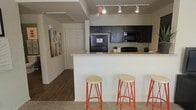 This single-story space showcases a thoughtfully designed open-concept layout where the kitchen flows naturally into the surrounding living areas. The kitchen is a standout feature, equipped with dark espresso-toned cabinetry that contrasts elegantly against light granite countertops. Black appliances, including a refrigerator and microwave, lend a cohesive, modern aesthetic throughout the cooking space. A peninsula-style breakfast bar extends from the kitchen, accompanied by three orange-accented bar stools that introduce a warm, vibrant touch to the neutral palette. Track lighting runs along the ceiling, providing focused illumination across the kitchen workspace. The flooring transitions from dark hardwood in the hallway area to carpet in the adjacent living space, clearly delineating functional zones while maintaining visual continuity. A corridor visible from the kitchen leads to additional rooms, with a bold piece of abstract wall art adding a decorative focal point along the neutral-toned walls. The overall ambiance is clean and practical, with a modern sensibility defined by the contrast of dark cabinetry, light surfaces, and warm accent details.