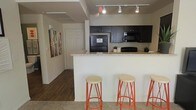 This single-story space showcases a thoughtfully designed open-concept layout where the kitchen flows naturally into the surrounding living areas. The kitchen is a standout feature, equipped with dark espresso-toned cabinetry that contrasts elegantly against light granite countertops. Black appliances, including a refrigerator and microwave, lend a cohesive, modern aesthetic throughout the cooking space. A peninsula-style breakfast bar extends from the kitchen, accompanied by three orange-accented bar stools that introduce a warm, vibrant touch to the neutral palette. Track lighting runs along the ceiling, providing focused illumination across the kitchen workspace. The flooring transitions from dark hardwood in the hallway area to carpet in the adjacent living space, clearly delineating functional zones while maintaining visual continuity. A corridor visible from the kitchen leads to additional rooms, with a bold piece of abstract wall art adding a decorative focal point along the neutral-toned walls. The overall ambiance is clean and practical, with a modern sensibility defined by the contrast of dark cabinetry, light surfaces, and warm accent details.