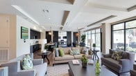 This well-appointed single-floor space showcases a thoughtfully designed open-concept layout that seamlessly blends comfort and sophistication. The main living area is anchored by a generously sized beige sofa adorned with colorful accent pillows, complemented by coordinating armchairs and a substantial dark coffee table, all set atop a patterned area rug. Architectural interest is provided by the striking tray ceiling with recessed lighting, which adds depth and dimension to the room.

The kitchen area, visible from the main lounge, features bold dark cabinetry, stainless steel appliances, and a breakfast bar, creating a sleek and functional culinary space. Pendant lighting with warm-toned fixtures hangs throughout, enhancing the inviting ambiance.

Large windows along one wall flood the interior with natural light, offering views of lush greenery outside and creating a bright, airy atmosphere. Decorative elements — including tall sculptural lamps, potted fiddle-leaf fig trees, and curated accent pieces — add layers of texture and visual interest throughout the space.

Sliding barn-style doors provide a stylish partition between areas, while the consistent use of a neutral palette accented with blues and greens ties the entire space together into a cohesive, welcoming environment.