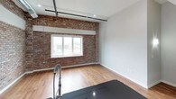 This single-floor loft showcases a compelling blend of industrial heritage and modern design. Exposed brick walls are a defining feature throughout the space, adding warmth and texture that contrast beautifully with the clean, neutral-toned walls and contemporary finishes. Hardwood flooring runs through the main living areas, tying the space together with a cohesive, polished look.

The kitchen is a standout area, featuring crisp white shaker cabinetry, dark granite countertops, and stainless steel appliances including a full-size refrigerator, range, and built-in microwave. A peninsula with a dark countertop provides additional prep and seating space, while an exposed brick accent wall and large window allow natural light to fill the room.

The bedroom is generously proportioned with soaring ceilings, exposed piping details, and a ceiling fan. A closet with built-in rods offers ample storage, and an adjoining full bathroom is accessible directly from the room. The bathroom features a sleek floating double vanity with an oversized mirror and light tile flooring, offering a clean, modern aesthetic.

A dedicated laundry area houses a stacked washer and dryer unit set against an exposed brick wall, adding both function and industrial charm. Exposed ductwork and black pipe accents throughout the space reinforce the loft's distinctive character.