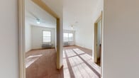 This single-floor residential unit offers a practical and well-organized layout across several interconnected rooms. The living areas and bedrooms feature warm-toned carpeting underfoot, while the kitchen and bathroom are finished with neutral ceramic tile flooring for easy maintenance. Each carpeted room is equipped with a ceiling fan and multiple windows dressed with horizontal blinds, allowing natural light to pour in throughout the day and creating a bright, airy atmosphere. Wall-mounted HVAC units provide individual climate control in the main rooms.

The kitchen is thoughtfully appointed with honey-toned wood cabinetry, dark granite-style countertops, a classic subway tile backsplash, a stainless steel sink, and white appliances including a range and over-the-range microwave. A hallway with recessed lighting connects the main living areas to the kitchen, maintaining an open and accessible flow throughout the unit.

The bathroom features matching light wood cabinetry with a dark countertop vanity, a tiled floor, and a full shower with a curtain enclosure. Additional doors off the main carpeted area provide access to a closet and the bathroom, keeping the layout organized and efficient. Overall, the space presents a clean, functional interior with consistent finishes and generous natural light throughout.