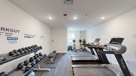 Located at 2140 Lakewood Rd in Toms River, NJ, this fitness room offers a compact yet efficiently organized exercise space designed to support a well-rounded workout routine. The room features a clean, modern aesthetic with crisp white walls adorned with motivational fitness-themed typography, adding an energizing atmosphere to the space. Flooring consists of durable, striped carpet tiles that provide both comfort and traction during exercise. Recessed lighting throughout ensures the room is evenly and adequately illuminated. The cardio section is equipped with multiple treadmills and an upright stationary bike, all positioned to allow comfortable use. A dedicated free weights area includes a two-tier dumbbell rack with a range of weights, a weight bench, and a stability ball, offering versatility for strength training. Potted plants add a touch of greenery, softening the utilitarian feel of the space. A window on one wall allows for a degree of natural light. Safety features include a fire extinguisher and fire suppression system. The layout is thoughtfully arranged to maximize the usable floor area, making it a functional and welcoming environment for residents seeking convenient on-site fitness facilities.