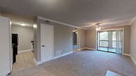 This single-floor residence is designed with a practical, open layout that connects the main living and dining areas seamlessly. The living room is carpeted in a neutral tone and benefits from a ceiling fan with a light fixture, crown molding, and a sliding glass door that opens to an outdoor balcony, allowing natural light to filter into the space. An adjacent bedroom mirrors this comfortable aesthetic, featuring the same plush carpet, a ceiling fan, and a window that provides additional natural light and outdoor views.

The galley-style kitchen is efficiently arranged with white shaker cabinetry lining both sides, dark granite-style countertops, and light wood-look flooring. Black appliances — including a refrigerator, electric range, and dishwasher — are integrated throughout, providing a functional contrast to the white cabinetry. The kitchen connects conveniently to the main living area.

The bathroom is clean and well-appointed, featuring a tub-shower combination with built-in shelving, a toilet, and light tile flooring. Throughout the unit, consistent use of gray-toned walls, white trim, and textured ceilings creates a cohesive, understated ambiance.