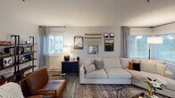This single-floor residence offers a thoughtfully designed layout encompassing a living room, kitchen, two bedrooms, and two full bathrooms. The living room serves as a welcoming centerpiece, featuring multiple windows with patterned curtains that allow generous natural light to fill the space. Neutral gray walls, textured ceiling, and plush carpeting provide a warm, cohesive backdrop throughout.

The kitchen is equipped with white cabinetry, white countertops, stainless steel appliances including a range, microwave, and refrigerator, and light wood-tone flooring that adds a clean, modern feel.

The primary bedroom is generously proportioned, offering ample room for a seating area alongside the main sleeping space, with a window overlooking greenery and a wall-mounted air conditioning unit. A second bedroom features a sliding closet and comfortable carpet flooring. Both rooms are finished with soft gray walls and textured ceilings.

The two bathrooms are consistent in style, each featuring a tub-shower combination, white vanity with modern fixtures, wood-tone flooring, and a large mirror. The overall aesthetic throughout the residence is contemporary and neutral, offering a versatile and comfortable living environment.