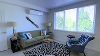 This single-floor space features a thoughtfully designed living area that balances modern functionality with eclectic character. The room is anchored by a sage green mid-century modern sofa adorned with colorful accent pillows, complemented by a classic blue armchair that adds a traditional counterpoint. A round wooden coffee table sits atop a bold black-and-white geometric area rug, creating a defined and inviting seating arrangement. A wall-mounted mini-split air conditioning unit ensures year-round climate comfort, while a distinctive wooden propeller mounted on the wall serves as a striking decorative focal point. Natural light fills the room through a large window fitted with white horizontal blinds, offering views of lush green foliage outside. The clean white walls and light-toned flooring enhance the brightness of the space. A floor lamp provides warm ambient lighting, and a small side table adds practical convenience to the seating area. The overall ambiance is casual yet curated, blending contemporary and vintage elements to create a welcoming, lived-in atmosphere.