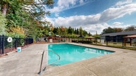Located at 970 Windy Hill Rd SE in Smyrna, Georgia, this outdoor community amenity space centers around a generously sized swimming pool with clear, turquoise water. The pool is framed by a wide concrete deck that provides ample room for lounging and socializing. Safety features are prominently in place, including pool markers and posted pool rules signage along the perimeter fence.

The area is enclosed by a classic black wrought-iron fence, offering both security and an open, airy feel. Within the enclosure, a covered gazebo with a dark umbrella canopy provides a shaded retreat for relaxing out of the sun. Seating with umbrella-shaded tables is also available along the deck perimeter.

A children's playground equipped with slides and climbing structures is situated just beyond the pool fence, making this a family-friendly destination. Mature trees, including tall pines and deciduous varieties displaying seasonal color, surround the space and provide a natural, park-like backdrop.

A utility building adjacent to the pool houses additional facilities, and landscaped plantings with ornamental grasses and shrubs add greenery throughout. The overall ambiance is welcoming and well-maintained, offering a comfortable outdoor retreat set against the backdrop of a residential community.