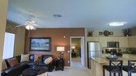 This single-floor residence showcases a thoughtfully designed open-concept layout that seamlessly connects the living and kitchen areas. The living room is anchored by a rich brown accent wall, which provides a warm, inviting contrast to the neutral surrounding walls and light-colored carpeted flooring. A ceiling fan with a light fixture overhead ensures both comfort and illumination throughout the space. The living area is further enhanced by a framed landscape painting, a leather sofa with decorative pillows, and coordinating dark wood accent tables, contributing to a cohesive, comfortable aesthetic.

The kitchen is directly accessible from the living area and features a stainless steel refrigerator, light-toned cabinetry, and a countertop bar with seating — ideal for casual dining or entertaining. The open sightlines between both spaces create a sense of generous square footage and ease of movement.

A doorway visible from the living room leads to an additional room, suggesting functional separation of private and common spaces. The overall ambiance is warm and practical, combining earthy tones with modern finishes for a balanced, livable environment.