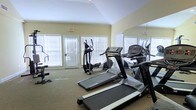 This fitness center is thoughtfully designed to support an active lifestyle in a bright and welcoming environment. The space is outfitted with a selection of cardio equipment, including multiple treadmills and an elliptical trainer, providing users with a variety of workout options. Large windows fitted with horizontal blinds line the walls, allowing generous amounts of natural light to fill the room while maintaining privacy. A full-length wall mirror spans one side of the space, visually expanding the room and allowing users to monitor their form during exercise. The vaulted ceiling adds an open, airy quality to the interior, enhancing the sense of spaciousness. Neutral-toned walls and carpeted flooring contribute to a clean, comfortable atmosphere. An exit door provides convenient access to the outdoors, adding to the functional layout of the space. The overall design balances practicality with comfort, making it an inviting facility for daily fitness routines.