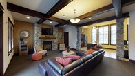 This inviting community lounge is designed to offer both comfort and style in a single, well-appointed space. The focal point of the room is a striking floor-to-ceiling stacked stone fireplace flanked by matching stone columns, which lend a warm, lodge-inspired aesthetic to the interior. Above, dark-stained exposed wood beams frame a coffered ceiling, adding architectural depth and a sense of craftsmanship throughout. Recessed lighting and a central pendant fixture ensure the space is well-lit, complementing the natural light that filters in through slatted windows along the far wall. The seating area is generously furnished with a large leather sectional sofa accented with colorful throw pillows, paired with additional armchairs and a round accent table, creating an ideal gathering environment. The open layout flows seamlessly into an adjacent kitchenette, which features dark cabinetry, a stainless steel refrigerator, and a counter with bar seating — perfect for casual entertaining. Warm earth-toned walls in shades of tan and yellow enhance the overall ambiance, making this a welcoming and functional communal space suited for relaxation and social interaction.