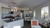This single-floor space showcases a thoughtfully designed open-concept layout that integrates the kitchen, dining, and living areas into one cohesive environment. The kitchen is a standout feature, outfitted with white cabinetry, dark countertops, and stainless steel appliances, including a full-sized refrigerator. A central island with an undermount sink provides additional workspace and casual seating. Overhead, a series of stylish pendant lights with decorative shades adds visual interest and focused illumination. Upper cabinetry includes a distinctive diamond-pattern wine rack, combining function with design detail. Recessed lighting throughout ensures the space remains bright and well-lit. The dining area accommodates a glass-top table with white chairs, lit by a drum-style pendant fixture. Natural light filters in through a nearby window with horizontal blinds. Dark hardwood-style flooring runs throughout, grounding the light color palette of white walls and cabinetry. The living area, partially visible, features contemporary white seating accented with an orange throw pillow, adding a warm contrast to the otherwise neutral tones. The overall ambiance is clean, modern, and inviting.