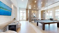 Located at Sawgrass Bay in Clermont, Florida, this elegantly designed single-floor clubhouse offers a generous, open-concept layout filled with natural light and contemporary architectural details. Soaring ceilings and floor-to-ceiling windows define the main living and dining areas, flooding the space with sunlight and providing views of the surrounding community. Light wood-look tile flooring runs throughout, complementing a warm, neutral color palette.

The living area features a curated seating arrangement, a built-in display wall with framed artwork, and a striking textured stone accent wall housing a large flat-screen television. Adjacent, the game room is anchored by a full-size pool table and a shuffleboard table, set beneath a rich wood-paneled ceiling with recessed and track lighting that adds warmth and depth.

The dining area is anchored by a round white table with cane-back chairs, bathed in light from expansive multi-pane windows framed by sheer draping curtains. A vertical wood slat partition elegantly separates spaces while maintaining an airy, open feel, and a kitchen bar area is visible beyond.

The entry corridor features glass-paneled doors, a textured accent wall, and a floating wood shelf, creating a polished and welcoming arrival experience. Throughout, the design strikes a balance between refined and relaxed — ideal for gathering and community living.