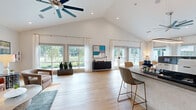 This thoughtfully designed single-floor community space combines an airy, open-concept layout with modern finishes and an abundance of natural light. The vaulted ceilings, accented by ceiling fans, lend a grand sense of height and openness throughout. Light-toned hardwood flooring runs seamlessly across the entire space, enhancing the clean, contemporary aesthetic.

The living area is generously proportioned, featuring large picture windows and glass French doors that frame lush outdoor views, including a serene pond and surrounding greenery. Sheer white curtains frame the windows, diffusing natural light while maintaining a bright, welcoming atmosphere. A seating area with a leather sofa and accent chairs provides a comfortable gathering spot, while a tasteful piece of abstract wall art adds a pop of color.

The main kitchen is well-appointed with gray shaker-style cabinetry, dark countertops, stainless steel appliances including a French door refrigerator, and a spacious peninsula with bar seating — ideal for casual dining or socializing. A striking oval pendant light anchors the kitchen visually. A secondary kitchenette, featuring a sink, dishwasher, and herringbone tile backsplash, offers additional utility. Glass double doors connect the space to adjacent areas, making it well-suited for community or event use.