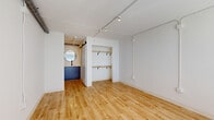 Located at 2306 Brick Church Pike in Nashville, TN, this efficiently designed single-floor unit offers a clean, modern aesthetic across a living room and bathroom. The main living area features warm light wood flooring that extends throughout, creating a cohesive and inviting atmosphere. Track lighting runs along the ceiling, providing flexible, directional illumination, while exposed conduit adds an industrial touch. An open built-in closet with double wooden shelving rods offers practical storage directly within the living space. A window fitted with horizontal blinds allows natural light to filter in, complemented by a wall-mounted HVAC unit for year-round comfort. Visible ductwork along the ceiling adds to the loft-inspired character of the space. Adjacent to the main room, a compact kitchenette is visible through an open doorway, equipped with navy blue cabinetry and a microwave, maintaining the bold, modern color palette. The bathroom is accessed via a sliding barn door and features a round wall mirror, navy vanity cabinetry with a black faucet, and textured wall accents. A dedicated walk-in shower is finished with clean white tile and fitted with a matte black shower valve, lending a contemporary, refined finish to the space.