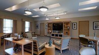 Located at 300 Valley Heights Drive in Williamsport, Pennsylvania, this single-floor space offers a warm and inviting atmosphere designed for comfort and community. The main common area serves as a central gathering space, highlighted by a series of decorative pendant lights suspended from a ceiling accented with recessed skylight panels that invite natural light into the room. A large, built-in wooden entertainment center anchors the space, housing a flat-screen television alongside open shelving displaying an array of curated decorative items. Comfortable armchairs with light wood frames and upholstered cushions are arranged throughout, encouraging relaxed conversation and leisure. The patterned carpet adds a sense of coziness underfoot, while warm-toned wood baseboards and trim complement the neutral wall palette. Additional seating and workspaces are thoughtfully positioned near the windows, which allow natural light to filter in through sheer curtains. Framed artwork adorns the walls, contributing to the refined yet approachable ambiance. The overall design balances functionality with aesthetic appeal, creating a communal environment that feels both purposeful and comfortable for its occupants.