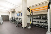 This fitness center offers a comprehensive workout environment designed with both functionality and modern aesthetics in mind. The space features a clean industrial character, defined by exposed concrete columns, crisp white walls, and durable dark rubber flooring throughout. Linear LED lighting runs across the ceiling, providing bright, even illumination across the entire floor.

The cardio zone is outfitted with multiple treadmills and elliptical trainers, each positioned in front of wall-mounted flat-screen televisions for an engaging workout experience. A dedicated strength training area includes a multi-function cable machine, a weight bench, and a dumbbell rack with a wall mirror, catering to a full range of resistance exercises. A standout feature is the Freedom Climber climbing wall, which adds a dynamic and unique training element to the space.

An open flex area with clean white walls and the same rubber flooring provides versatile room for stretching, bodyweight exercises, or group fitness activities. A window positioned near the entry door brings in a hint of natural light and a glimpse of greenery from outside. The overall ambiance is sleek, purposeful, and motivating — a well-rounded facility suited for a variety of fitness routines.
