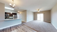 This single-floor space showcases an open-concept layout that seamlessly connects the kitchen and living areas. The kitchen features dark cabinetry, stainless steel appliances, granite countertops with a breakfast bar overhang, and pendant lighting that adds a polished touch. A decorative chandelier near the entry enhances the welcoming atmosphere.

The transition from the kitchen into the living area is marked by a shift from hardwood-style flooring to plush wall-to-wall carpeting, creating a distinct yet cohesive flow between spaces. Neutral beige walls provide a versatile backdrop throughout. The living area is generously sized, highlighted by a ceiling fan with integrated lighting that promotes comfort and airflow.

A sliding glass door at the far end of the living area opens to a private balcony, allowing natural light to fill the space and offering outdoor access. The overall ambiance is clean, modern, and functional, with thoughtful finishes that balance practicality and style throughout the open layout.