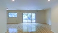 This single-floor space presents a clean, open layout designed to maximize natural light and a sense of spaciousness. The main living area is anchored by polished hardwood floors that reflect light beautifully, lending warmth and a classic elegance to the interior. A set of expansive sliding glass doors serves as a focal point along one wall, flooding the room with natural light and providing direct access to an outdoor deck. The deck overlooks a lush, tree-lined landscape, offering a serene and private natural backdrop. A secondary window to the side further enhances the brightness of the space, ensuring that natural light reaches every corner. The smooth, white ceiling features subtle recessed architectural detailing and integrated ventilation, contributing to the room's polished, well-maintained appearance. The neutral wall tones complement the warm tones of the hardwood flooring, creating a cohesive and inviting atmosphere. The open floor plan provides exceptional flexibility, making the space well-suited for a variety of uses. Overall, the combination of abundant natural light, quality hardwood flooring, and seamless indoor-outdoor connectivity gives this space a bright, welcoming, and versatile character.