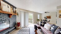 Located at 6025 N Jefferson St in Kansas City, MO, this single-floor home combines modern updates with warm, inviting character. The living room serves as the heart of the space, anchored by a striking dark-painted brick fireplace with a rustic wood mantle. Light wood-style flooring flows throughout the main living areas, complementing the neutral wall tones. Sliding glass doors open to an outdoor deck, allowing natural light to pour in while providing seamless indoor-outdoor access with views of surrounding greenery. A ceiling fan overhead and an adjacent dining area complete the open living and entertaining space.

The kitchen features crisp white cabinetry with matte black hardware, dark granite-style countertops, a white subway tile backsplash, and a full suite of stainless steel appliances including a range, dishwasher, and refrigerator. The bedrooms offer comfortable, carpeted retreats with natural light from window blinds and ceiling fans for added comfort. The bathrooms are clean and functional, featuring white cabinetry, dark countertops, and light wood-style flooring consistent with the rest of the home. The overall aesthetic is cohesive and modern with warm, bohemian-inspired touches throughout.