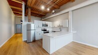 Located at 425 W Lakeside Ave in Cleveland's vibrant Lakeside neighborhood, this one-bedroom, one-bathroom loft-style unit masterfully blends industrial character with modern finishes. The open-concept kitchen and living area serves as the heart of the space, showcasing soaring exposed wood beam ceilings supported by original timber columns — a striking nod to the building's historic origins. Light hardwood flooring runs throughout, complementing the whitewashed brick walls that add texture and warmth. Large arched and rectangular windows flood the living area with abundant natural light.

The kitchen is well-appointed with white shaker cabinetry, a white quartz peninsula with bar seating, subway tile backsplash, and a full suite of stainless steel appliances. An in-unit washer is conveniently integrated into the kitchen island cabinetry. A hallway with exposed beam ceiling detail leads to the private bedroom, which features windows along one wall and continued hardwood flooring. Two closets provide ample storage. The bathroom includes a full tub/shower combination with clean, modern fixtures. The overall ambiance is a refined industrial aesthetic — open, airy, and full of character unique to Cleveland's adaptive reuse architecture.