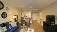 This single-floor residence offers a thoughtfully designed open-concept layout that seamlessly integrates the living, dining, and kitchen areas into one cohesive and inviting space. The living room features warm, textured accent walls and is anchored by a plush navy blue sofa, complemented by soft recessed lighting and crown molding that adds a refined architectural touch. Hardwood-style flooring flows continuously throughout, lending warmth and visual continuity across the entire floor plan. The dining area sits at the heart of the space, accommodating a round table with upholstered chairs, creating an intimate setting ideal for everyday meals or entertaining. The kitchen is bright and well-appointed, showcasing white cabinetry, a dark-toned countertop with a breakfast bar overhang, and stainless steel appliances including a large refrigerator. Recessed lighting illuminates the kitchen efficiently, while clean white finishes keep the space feeling open and airy. A hallway extends from the main living area, leading to additional rooms and featuring arched doorway details that add architectural character. The overall ambiance is warm, welcoming, and polished, with a neutral color palette punctuated by tasteful décor accents throughout.