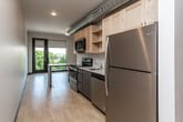 This well-appointed single-floor residence offers a thoughtfully designed open-concept layout that seamlessly connects the kitchen, dining, and living areas. Light hardwood flooring flows throughout the space, lending warmth and continuity to every room.

The kitchen is a standout feature, equipped with a full suite of stainless steel appliances including a refrigerator, electric range with over-the-range microwave, and dishwasher. Light maple cabinetry provides generous storage, while a built-in breakfast bar extends from the kitchen peninsula, offering a casual dining option. An exposed silver ductwork detail adds an industrial-modern character to the open ceiling above.

The living area is bathed in natural light, thanks to expansive floor-to-ceiling sliding glass doors that open onto a private balcony. The balcony offers scenic views of lush treetops and a city skyline, making it a serene outdoor retreat. Roller shades on the large windows allow for adjustable light control.

The bedroom is generously proportioned and includes a built-in closet with shelving and hanging rods for ample storage. The bathroom features a clean, minimalist design with a glass-enclosed walk-in shower, a floating vanity with an integrated sink, and wood-toned cabinetry. A thermostat in the hallway ensures comfortable climate control throughout.