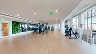 This professionally equipped fitness facility offers a comprehensive workout environment spread across a generously sized single floor. The space is defined by its bright, contemporary aesthetic, featuring light hardwood flooring, crisp white walls, and soaring double-height ceilings in several areas that create an open, energizing atmosphere.

The main strength training floor is lined with large mirrors and houses a full suite of cable machines, free weights, and resistance equipment, complemented by recessed lighting and a live green wall accent. A dedicated functional training zone features Octagon power racks, battle ropes, TRX suspension systems, and medicine balls, set on durable carpet flooring.

The cardio area is positioned along floor-to-ceiling glass windows, offering abundant natural light and outdoor views, and is equipped with ellipticals, stationary bikes, rowing machines, and an Assault Fitness rower. A separate Pilates studio benefits from the same dramatic glazed walls, housing multiple reformer machines and stability balls in a calm, sunlit setting.

A group exercise and studio zone features a Wellbeats interactive display screen, step platforms, and open floor space ideal for classes. A boxing station with a heavy bag and spinning bikes rounds out the facility's diverse training offerings. The overall design balances functionality with an upscale, welcoming ambiance.