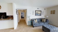 This single-floor residence offers a comfortable and functional layout designed to maximize living space. The living area serves as the heart of the home, featuring plush wall-to-wall carpeting that adds warmth underfoot, along with crisp white walls that reflect natural and ambient light throughout. Decorative wall sconces and framed artwork lend a polished, curated feel to the space.

A wide hallway extends from the living area, providing seamless access to the private quarters of the home, including a bedroom visible at the end of the corridor. The hallway is well-lit with overhead lighting and features a closet, offering practical storage solutions. Ceiling vents are present throughout, indicating a central climate control system for year-round comfort.

The open flow between the living space and the rest of the home creates a sense of continuity and ease of movement. The neutral color palette — soft beige tones on the floors paired with bright white walls and ceilings — gives the space a clean, airy aesthetic that is both inviting and versatile. Textured ceilings add a subtle architectural detail that complements the overall design. This thoughtfully laid-out residence balances everyday functionality with a welcoming ambiance.