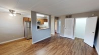This single-floor space offers a clean, functional layout designed for comfortable everyday living. The open-concept living area is anchored by warm hardwood-style flooring that extends throughout, creating a cohesive and inviting atmosphere. A defining architectural feature is the pass-through kitchen window, which connects the kitchen to the main living area, enhancing the sense of openness while maintaining a defined cooking space. The kitchen is equipped with stainless steel appliances — including a full-size refrigerator and microwave — complemented by wood-toned cabinetry and a stone-finish countertop. Neutral gray walls throughout the space provide a versatile backdrop suited to a variety of interior styles. A hallway visible from the main living area leads to additional rooms, offering a practical and well-organized flow. The space also features a louvered door, adding a classic architectural touch, along with ceiling ventilation to support airflow and comfort. Overall, the layout balances open communal areas with defined functional zones, making it a practical and well-finished space suitable for a range of occupants.