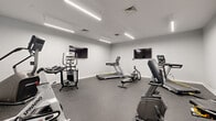 Located at 400 N Main St in Glen Ellyn, Illinois, this fitness center offers a clean, modern environment designed to support a comprehensive cardio-focused workout experience. The single-floor space features crisp white walls, a speckled rubber flooring ideal for high-traffic athletic use, and sleek linear LED lighting suspended from the ceiling, providing bright, even illumination throughout the room.

The facility is outfitted with a diverse selection of Matrix-brand cardio equipment, including treadmills, an elliptical trainer, a recumbent bike, a rowing machine, a spin bike, and a multi-station exercise machine. Multiple flat-screen televisions are mounted on the walls, allowing users to stay entertained or informed during their workouts. Sports-themed wall art — including vibrant cycling and running-inspired canvas prints — adds energy and visual interest to the space.

The layout is thoughtfully arranged to provide ample room between machines, promoting ease of movement and a comfortable workout environment. A frosted glass door provides access to and from the facility, lending a polished, professional aesthetic. The overall ambiance is contemporary and functional, reflecting a well-maintained, purpose-built fitness space suited for residents or members seeking convenient access to quality exercise equipment.
