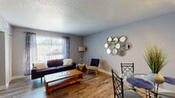 This single-floor residence offers a thoughtfully designed open-concept layout that seamlessly connects the living, dining, and kitchen areas. The living room is a welcoming space with warm hardwood-style flooring, a large window dressed with vertical blinds and patterned curtains that fill the room with natural light, and a decorative multi-circle mirror display as a standout wall feature. A wall-mounted flat-screen television anchors one end of the room, complemented by light gray flooring in that area. The dining space comfortably accommodates a glass-top table and chairs, positioned adjacent to the kitchen pass-through opening for easy entertaining. The galley-style kitchen features rich dark cabinetry, granite countertops, and stainless steel appliances including a microwave and dishwasher. The bedroom is a serene retreat with dark hardwood-style flooring, a ceiling fan with lighting, natural light from a window with blinds, and access to a spacious walk-in closet. The full bathroom is well-appointed with a granite-topped dark vanity, a framed mirror, and a tub-shower combination. Textured walls and ceilings are consistent throughout, adding a classic character to the overall design of this comfortable, move-in-ready space.