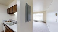 This single-story space features an open-concept layout that connects the kitchen and living areas seamlessly. The kitchen is equipped with a stainless steel sink, dark wood upper cabinetry, and an under-cabinet light fixture, providing functional workspace. A textured white column divides the kitchen from the living area and houses a wall-mounted electrical panel, offering convenient utility access. The living area is finished with neutral carpet and benefits from a large window that spans much of the exterior wall, allowing natural light to fill the space. A baseboard heating unit runs along the base of the window wall, providing efficient climate control. The ceilings are a standard height with a smooth white finish, and the overall color palette throughout remains light and neutral, contributing to a clean, practical atmosphere. The straightforward design and open flow between spaces make the layout both functional and easy to navigate.