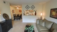 This single-story space showcases a thoughtfully designed open-concept layout that seamlessly connects the main living areas. The living room offers a comfortable, carpeted space anchored by a sofa and coffee table, with warm lighting provided by a classic table lamp. Decorative diamond-shaped wall art adds a stylish accent above the pass-through kitchen window, which serves as a functional and visually interesting architectural feature that links the living area to the kitchen.

The kitchen is accessible via the pass-through opening, which reveals wooden cabinetry, a light-toned countertop, and standard appliances including a refrigerator and range, providing an efficient and practical cooking space. The adjacent dining area accommodates a full dining set with multiple chairs, and features a ceiling fan with an integrated light fixture, ensuring comfortable airflow throughout the space. A decorative wall clock and framed artwork contribute to the warm, inviting ambiance of the dining zone.

Throughout the space, neutral wall tones and carpeted flooring create a cohesive and welcoming atmosphere suited for everyday living and entertaining.