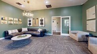 Located at 300 Gatehouse Ln in Odenton, MD, this single-level community amenity space offers a thoughtfully designed layout encompassing a fitness center, lounge, business center, and dining area. The exercise room is a standout feature, boasting soaring vaulted ceilings with warm wood paneling, recessed lighting, and large arched windows that flood the space with natural light. It is outfitted with a comprehensive array of cardio machines, strength equipment, free weights, and functional training gear, all set on durable rubber flooring. Adjacent to the fitness area, the inviting living room showcases sage-green walls, patterned carpet, and elevated decor including a leather sectional sofa, a sculptural chandelier, and curated artwork—creating a warm and social atmosphere. The hallway connects the spaces seamlessly, featuring a cozy seating nook with upholstered chairs and gallery-style wall art. The dining area doubles as a business center, highlighted by a textured stone accent wall, pendant lighting, iMac workstations along a built-in desk, a round concrete communal table with modern chairs, and wood-framed mirrors. Together, these spaces deliver a polished, contemporary environment designed for both productivity and relaxation.