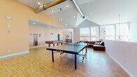 This single-floor community recreation space is thoughtfully designed to encourage social activity and relaxation. The main game area is the centerpiece of the room, boasting soaring vaulted ceilings with exposed industrial-style ductwork that adds a modern, loft-like character. Hardwood-style flooring runs throughout, unifying the open layout. A full-size ping-pong table occupies the generous central floor space, while a billiards table with a wall-mounted cue rack sits nearby, making the area well-suited for friendly competition.

Adjacent to the game area, a comfortable lounge zone features a large leather sectional sofa and a cluster of cushioned barrel chairs arranged around a coffee table. A flat-screen television is mounted on the warm peach-toned accent wall, and an expansive set of floor-to-ceiling windows floods the space with natural light while offering views of the surrounding neighborhood and mature trees, creating an inviting, open ambiance.

A separate dining nook provides a quieter retreat, furnished with a compact high-top table and chairs beneath decorative wall art, with easy access to an adjoining service area equipped with a small refrigerator and coffee station. Recessed lighting throughout ensures the space remains bright and welcoming at any hour.