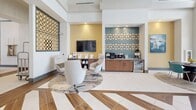 The Solaris Leasing Office in Kissimmee, Florida is a thoughtfully designed single-story space that blends professional functionality with inviting, contemporary aesthetics. Upon entry, soaring double-height ceilings and large floor-to-ceiling windows flood the reception area with abundant natural light, offering views of the surrounding greenery. The reception zone features a sleek leasing desk accented with teal tile detailing, paired with comfortable guest seating — all set against crisp white walls adorned with a striking gold geometric lattice panel that serves as a bold architectural focal point.

The lounge area is elegantly furnished with a plush sofa, curved accent chairs, and layered area rugs atop large-format porcelain tile floors inset with warm wood-toned chevron detailing. A curated art piece anchors the space, adding a creative touch to the refined interior.

Adjacent to the lounge, a well-appointed refreshment station with warm wood cabinetry, decorative tiled backsplash, and a beverage center invites guests to relax. The space opens into a generously sized amenity room featuring a pool table, bar-height seating, statement pendant lighting, and a large flat-screen TV — all beneath dramatic high ceilings that amplify the open, airy feel. The overall ambiance is modern, polished, and welcoming.