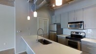 This single-floor space showcases a thoughtfully designed kitchen that blends modern and industrial aesthetics. The kitchen is anchored by a large island with a clean white countertop and an undermount sink fitted with a sleek, commercial-style spring faucet. Pendant lights hang above the island, providing focused task lighting that complements the exposed ceiling ductwork and metal conduit — signature industrial design elements that add character and visual height to the space.

The cabinetry is finished in a soft grey tone, offering a contemporary look that pairs well with the classic white subway tile backsplash lining the cooking area. Stainless steel appliances, including a refrigerator and an electric range with an overhead exhaust hood, provide a cohesive, professional-grade feel throughout the kitchen. Dark lower cabinetry creates a grounded contrast against the lighter upper cabinets and countertops.

A louvered door in the background hints at additional storage or utility space. Track lighting on the ceiling supplements the pendant fixtures, ensuring the kitchen is well-lit throughout. The overall ambiance is clean, functional, and stylishly modern.