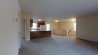 This single-floor space is designed with an open-concept layout that promotes a sense of spaciousness and ease of movement throughout. The kitchen is centrally positioned and features dark wood cabinetry, a white refrigerator, and a peninsula-style counter that provides both workspace and a natural divider between the kitchen and the adjoining living area. A sink is integrated into the peninsula, and recessed lighting overhead keeps the kitchen well-lit. The countertops offer ample prep space, and the cabinetry provides generous storage. The living area extends generously from the kitchen, finished with neutral wall-to-wall carpeting that adds warmth and comfort underfoot. Multiple windows are distributed throughout the space, allowing natural light to pour in and creating a bright, airy atmosphere. The walls are painted in a soft, neutral tone that complements the overall palette. A staircase railing is visible toward the back of the living area, indicating access to a lower level. The flush-mount light fixtures and recessed lighting ensure consistent illumination throughout. The open flow between the kitchen and living zones makes this space well-suited for both everyday living and casual entertaining.