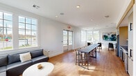 Located at 1001 NW Cernan Dr in Huntsville, Alabama, this single-floor communal space offers a thoughtfully designed open-plan layout that combines a living room, dining area, and kitchen into one cohesive environment. Warm-toned hardwood flooring runs throughout, complementing the crisp white walls and recessed lighting that keep the space feeling bright and welcoming.

The living area is anchored by a large L-shaped sectional sofa in charcoal gray, paired with a round white coffee table. Multiple oversized windows and a set of glass-paneled French doors flood the room with natural light while providing outdoor views of the surrounding neighborhood. A large abstract painting adds a vibrant touch of color to the corner seating nook.

The dining zone features a long, dark-topped communal bar table with high stools, ideal for collaborative or casual dining. Smaller round marble-top tables with modern chairs are tucked near the windows, creating intimate seating nooks bathed in sunlight.

The kitchen is finished with blue-gray countertops, a stainless steel undermount sink with a matte black faucet, white cabinetry, and a vertical white subway tile backsplash accented by warm gold sconce lighting. A hallway with a sliding panel door connects the space to an adjacent fitness area, rounding out this well-appointed community hub.